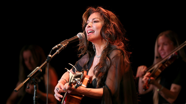 Shania Twain in an all-black outfit, singing on stage while playing guitar