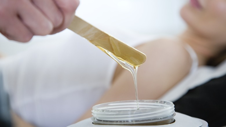 A waxer pulling a stick coated with wax out of a jar to apply to a female client