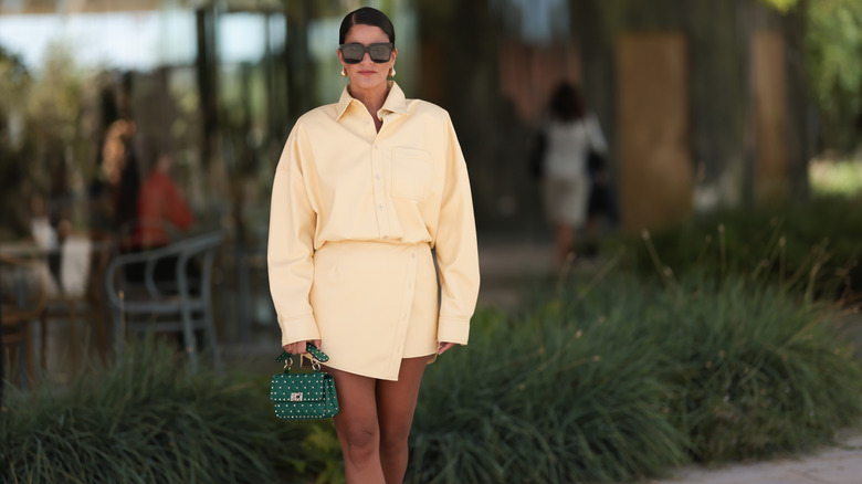A woman poses in a cream shirt and skirt outfit with a green bag