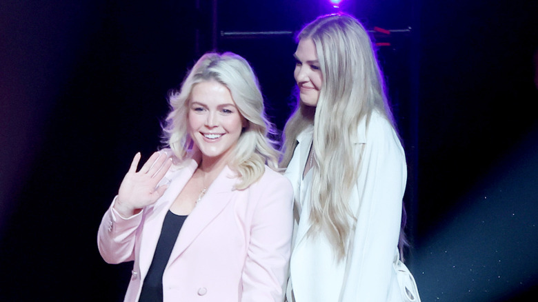 Erika Kirk and Karoline Leavitt walk onstage at a Turning Point USA event. The image is cut off at their waists; Kirk is a pale blonde woman wearing a white suit jacket. Leavitt is a pale blonde woman wearing a pink blazer and black tank top.