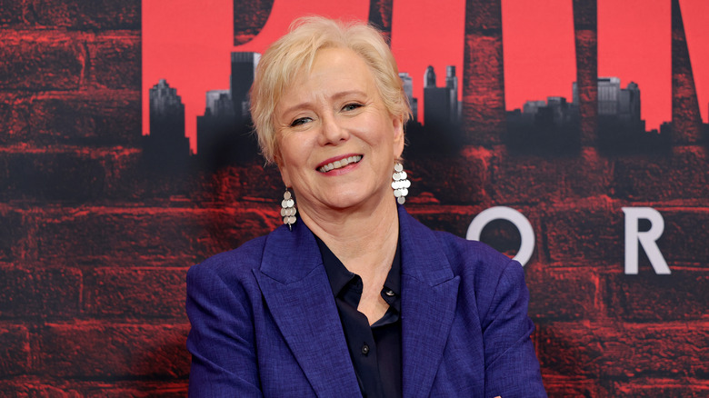 Eve Plumb wears a blue jacket and dangly earrings while posing in front of a red backdrop