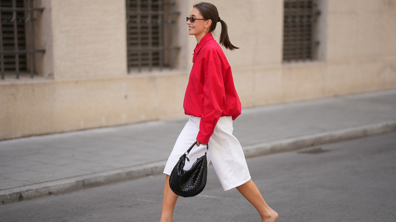 A woman crosses the street wearing a red jacket, white shorts, and carrying a black purse