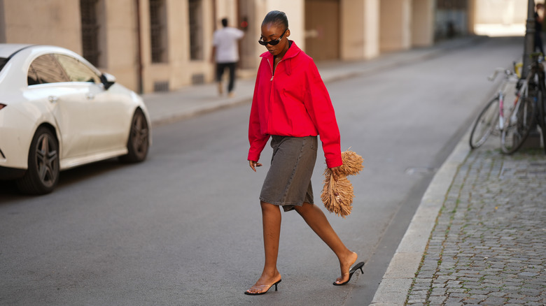 A woman crosses the street wearing a red jacket and denim skirt