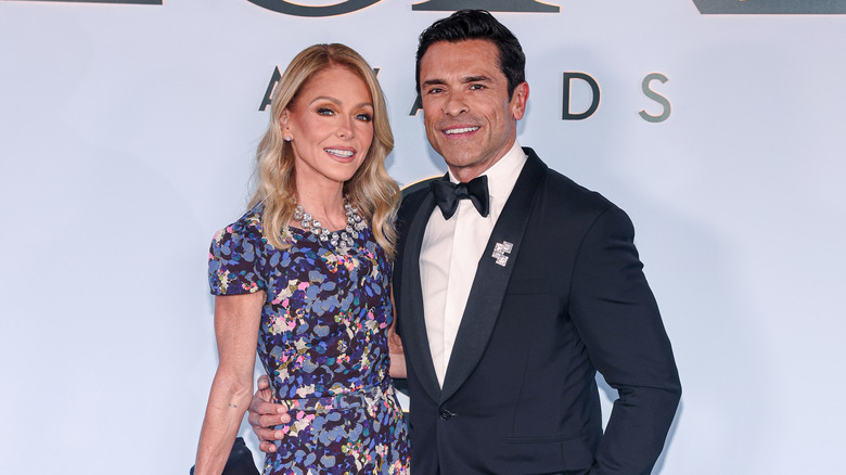 Kelly Ripa in a purple floral dress standing next to Mark Consuelos in a navy blue tuxedo.