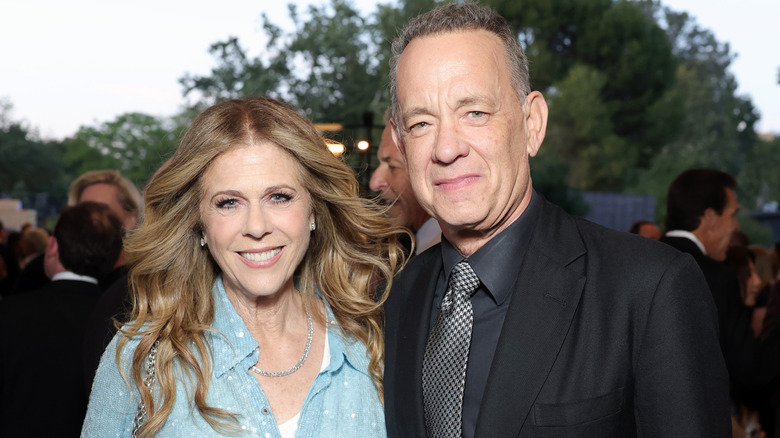 Rita Wilson in a baby blue top with her hair down standing next to Tom Hanks in a black suit.