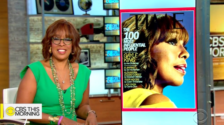 Gayle King smiling next to a photo of her TIME magazine cover wearing glasses, a green dress, and long green necklaces.