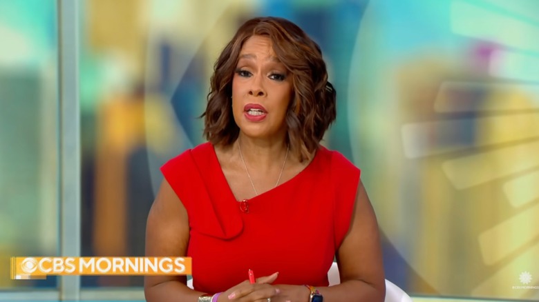 Gayle King wearing red lipstick and a red dress sitting at a news desk.