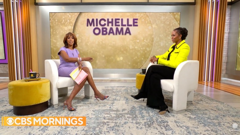 Gayle King wearing a lilac dress with her legs crossed interviewing Michelle Obama wearing black pants and a neon-yellow top.