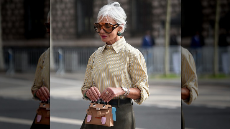 A woman with short gray hair wearing a striped collared shirt and sunglasses with gold and silver rings and a silver bracelet.