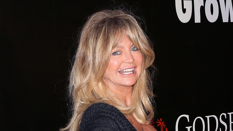 Goldie Hawn smiling at an event wearing a black top against a black background.