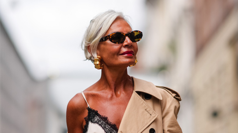 A woman poses wearing oval sunglasses and a beige trench coat hangs off her shoulder
