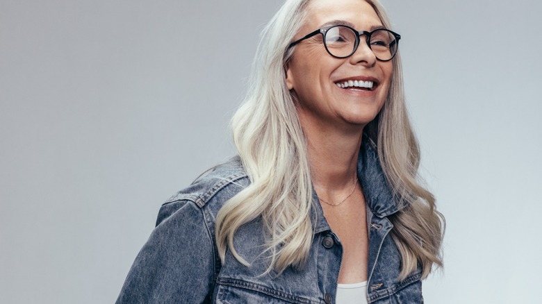 A woman smiling with long gray hair wearing glasses and a denim jacket.