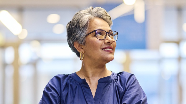 A woman with a dark-gray pixie cut wearing glasses and a blue top looking to the side.