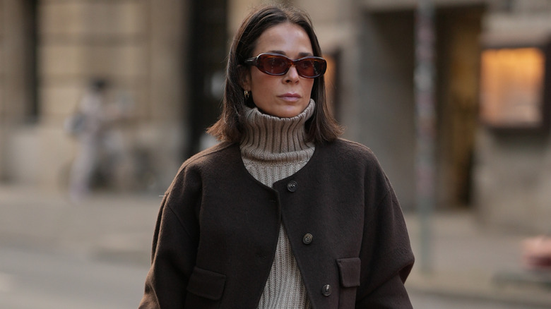 A woman wears sunglasses, a turtleneck, and brown jacket