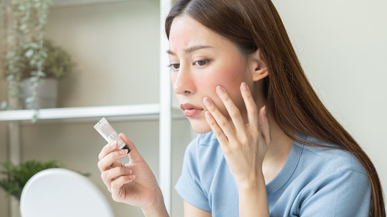 Woman holding cosmetic item and looking at skin in mirror