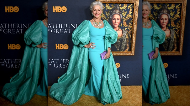 Helen Mirren posing in front of a painting in 2019 wearing an aqua dress with a poofy cape and statement jewelry.