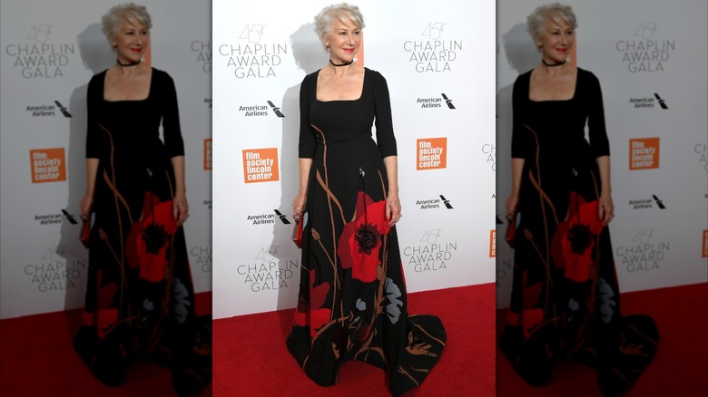 Helen Mirren posing on the red carpet in 2018 wearing a long black dress with red flowers.