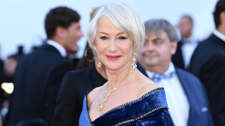 Helen Mirren smiling at a red carpet event in 2018 wearing a blue velvet dress and gold jewelry.