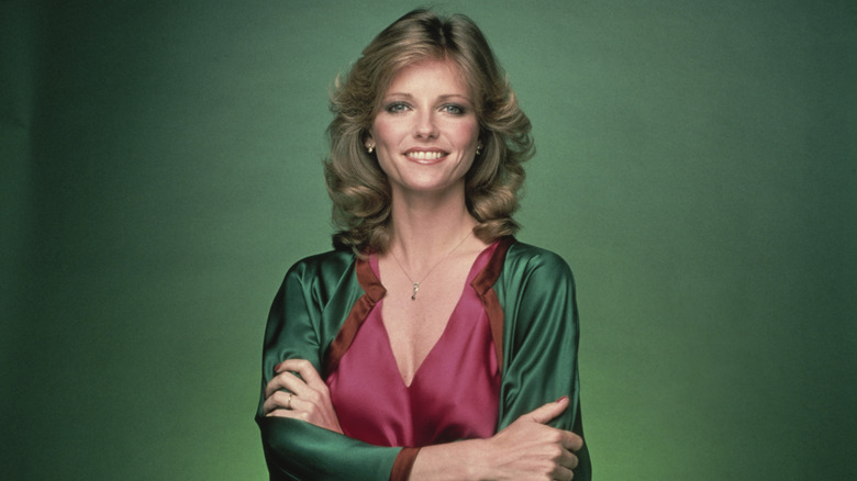 Model Cheryl Tiegs wearing a red satin top and green satin jacket with her arms crossed in front of a green background.