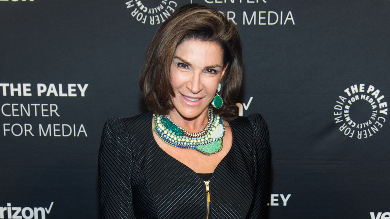 Hilary Farr posing at a red carpet event in a black dress and intricate green necklace and earrings.