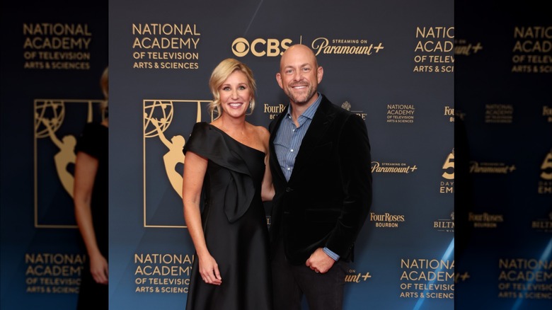 Jenny and Dave Marrs pose on the Emmy's red carpet. Jenny is wearing a one-shoulder black satin gown. Dave is wearing a black suit with a blue striped button down underneath.