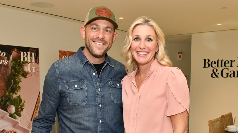 Jenny Marrs poses alongside husband Dave Marrs at a Better Homes & Garden's event. Dave is a fair-skinned man with close facial hair. He is wearing a green baseball hat and a blue denim shirt. Jenny is a fair-skinned woman with blonde hair. She is wearing a blush pink short-sleeved blouse and thin silver hoop earrings.