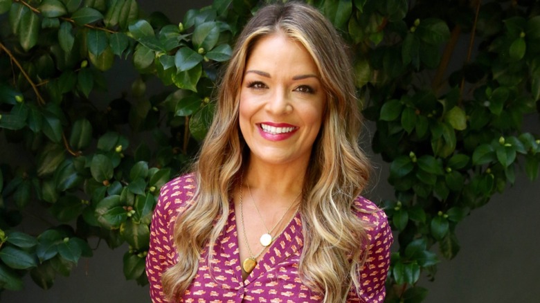 Sabrina Soto smiling in front of a leafy background wearing a pink and yellow top with multiple necklaces.