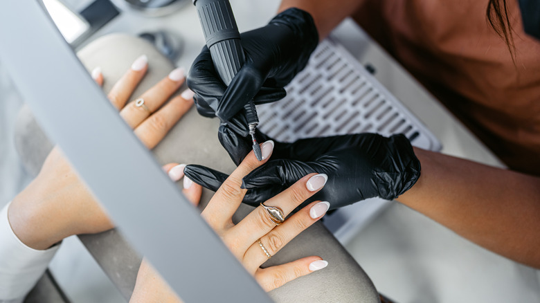Woman getting her french manicure acrylic nails buffed by a technician wearing black gloves at a salon.