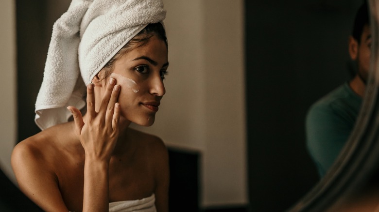 A woman in a towel applies lotion to her face.