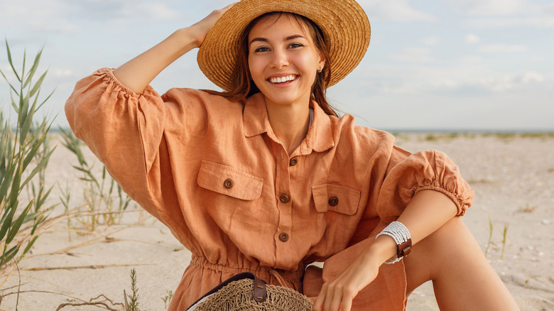woman in orange linen romper sits on the beach with a straw hat and wicker purse