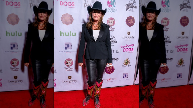 Actress Kelly LeBrock on the red carpet wearing a white shirt with a black jacket and pants with a black cowboy hat and red cowboy boots.