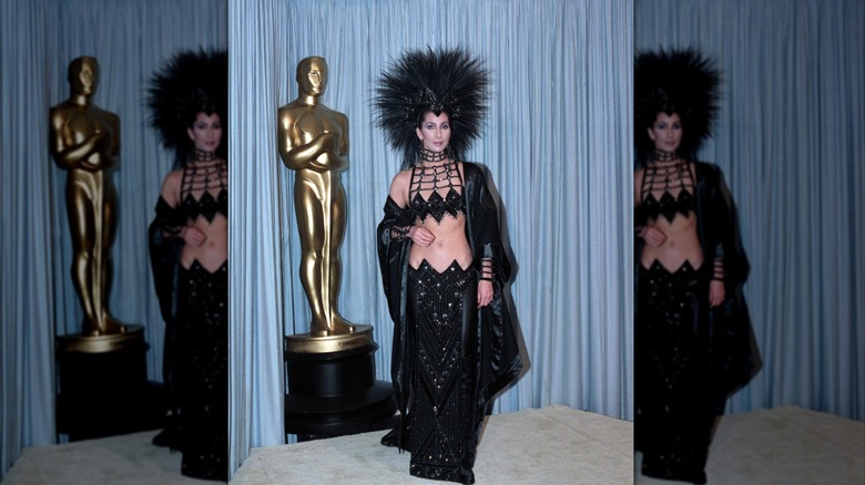 Cher wearing a glittery black outfit and large feathery black headdress in front of an award statue at the 1986 Oscars.