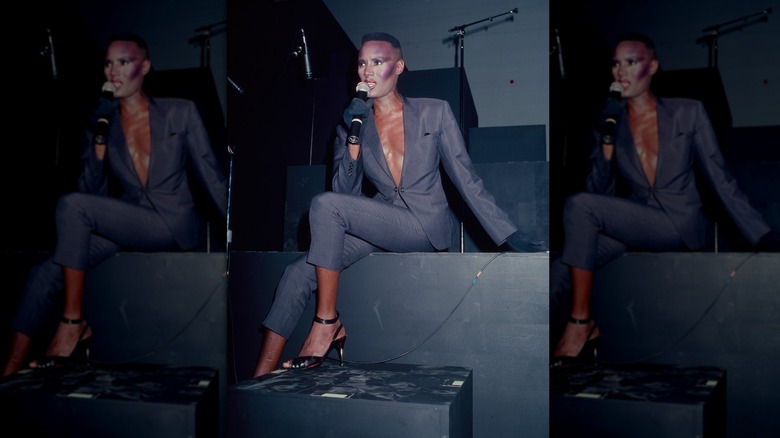 Grace Jones wearing a gray suit with a deep-V neck while sitting down and singing on stage.