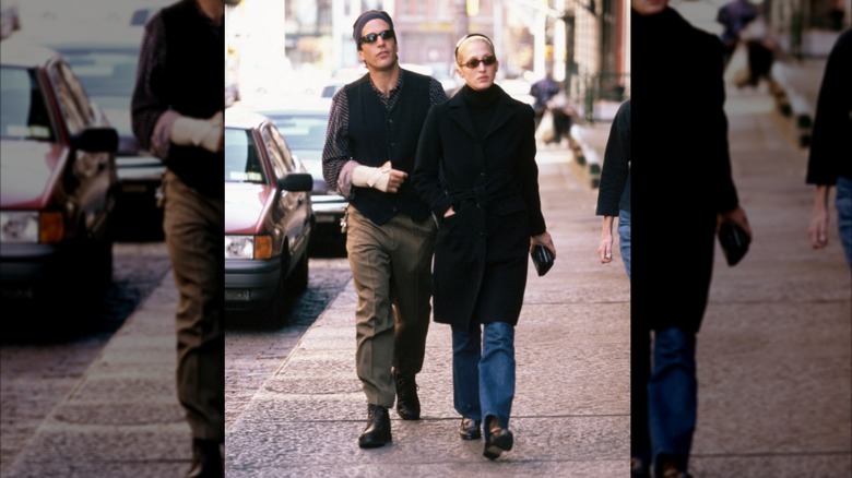 Carolyn Bessette-Kennedy in bootcut jeans, black trench coat, and heeled boots walking alongside JFK Jr.