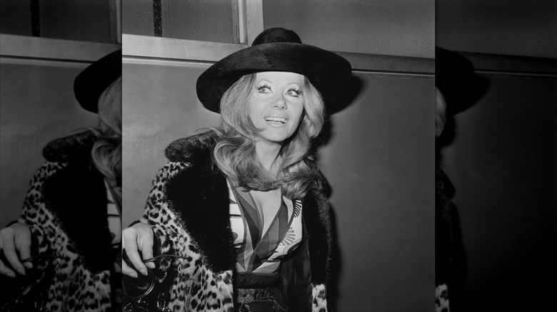 Ingrid Pitt smiles wearing a leopard-print jacket and a wide-brimmed hat