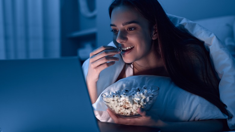 Woman snacking on popcorn with laptop in bed