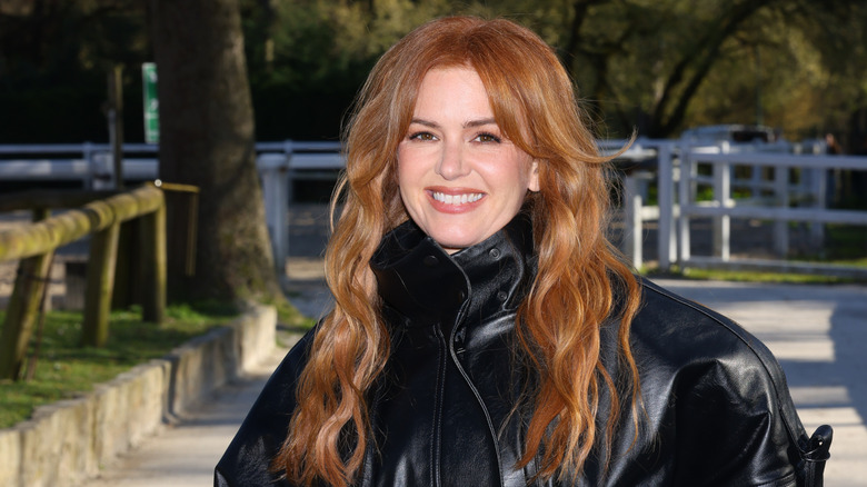 Isla Fisher smiling outside wearing a black leather jacket with her hair down.