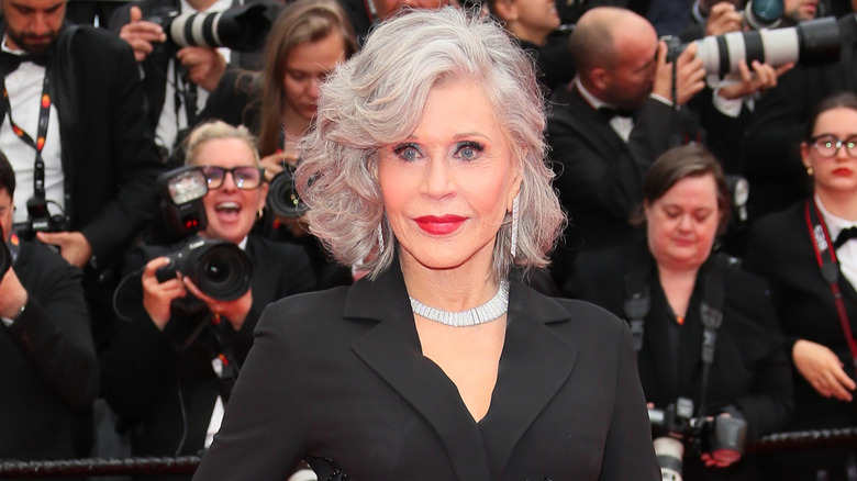 Jane Fonda posing at a red carpet event wearing red lipstick, a black jacket, and a diamond necklace.
