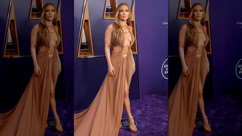 Jennifer Lopez on the red carpet wearing a long beige dress with cut-outs and metallic details.