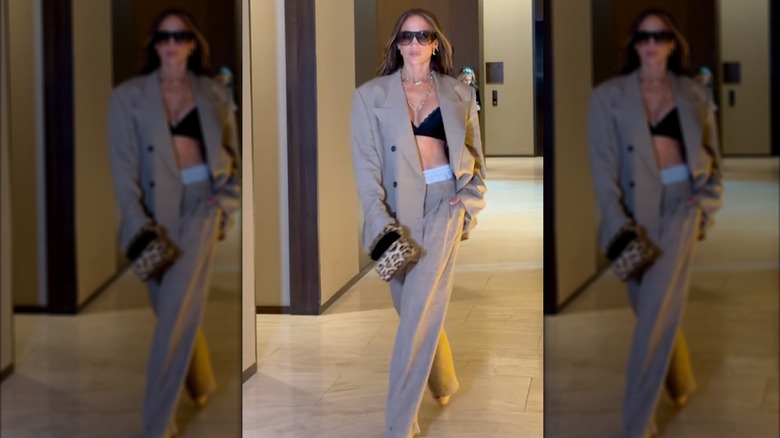 Jennifer Lopez walking and wearing a beige pants and jacket set with a black bra top, a leopard-print clutch, and sunglasses.
