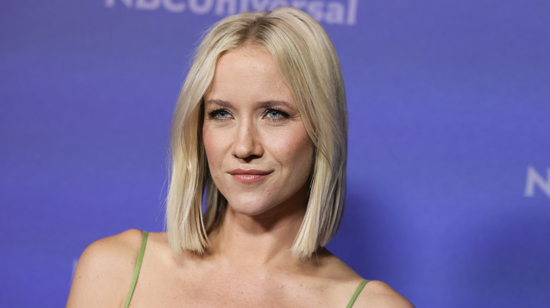 Blond Jessy Schram poses in front of an NBC Universal backdrop, wearing a green spaghetti strap dress