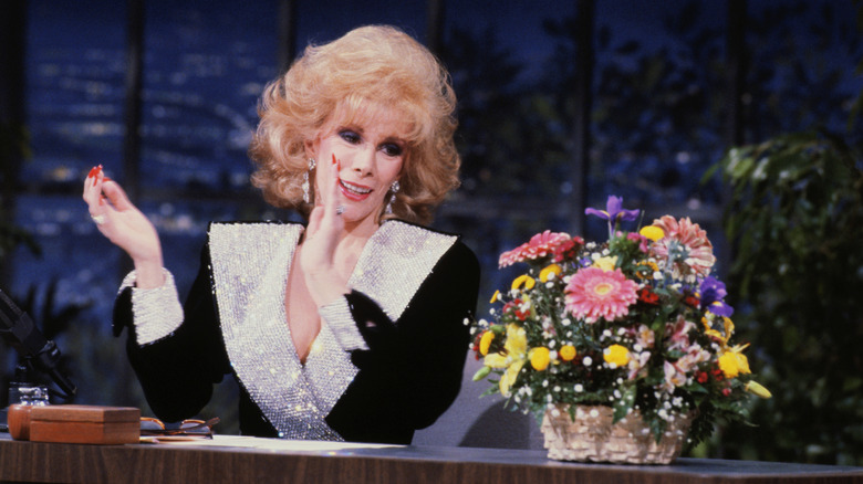 Joan Rivers claping her hands while looking at a bouquet of flowers on a late night show.