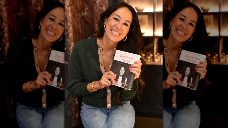 Joanna Gaines posing with her book wearing jeans and a dark green sweater.