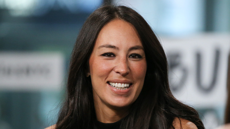 Joanna Gaines smiling with her hair down in a black tank top against a blurry background.