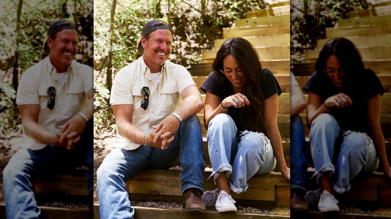 Chip Gaines wearing a white shirt and jeans with a backwards cap and Joanna Gaines wearing jeans and a black tee laughing together sitting on stairs.