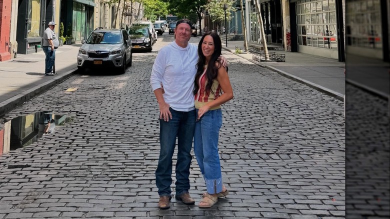 Chip Gaines in jeans and a T-shirt and Joanna Gaines wearing a tank top and jeans pose with their arms around each other.