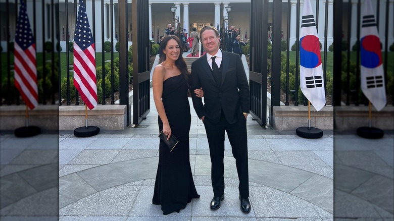 Chip Gaines in a suit and Joanna Gaines wearing a strapless black gown posing in front of flags.