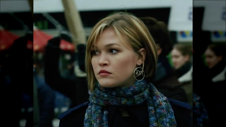 Julia Stiles as Nicky in "The Bourne Supremacy," wearing a a tailored purple jacket, statement earrings, and a colorful skinny scarf.