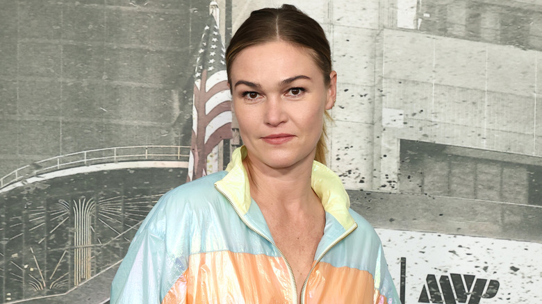 Julia Stiles smiles wearing a pastel yellow, blue, and orange bomber jacket.