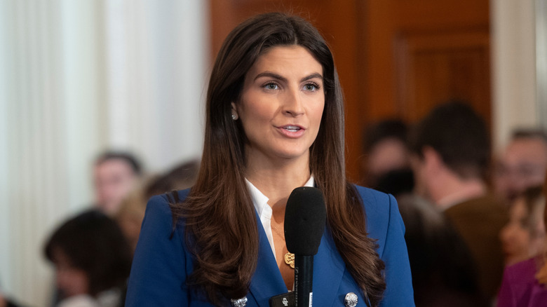 Kaitlan Collins speaks into a microphone while wearing a royal blue blazer and a gold pendant necklace.